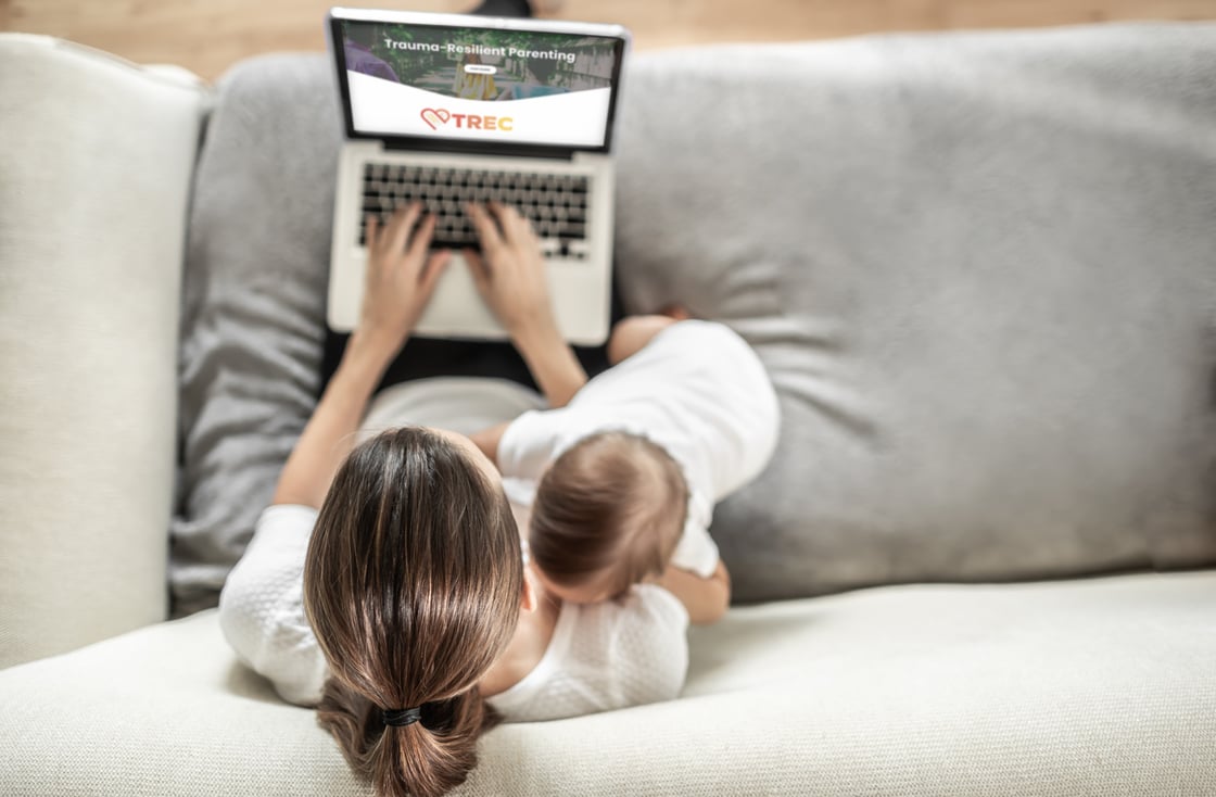 Mom sitting on sofa using laptop with baby crawling on her arm while she works.