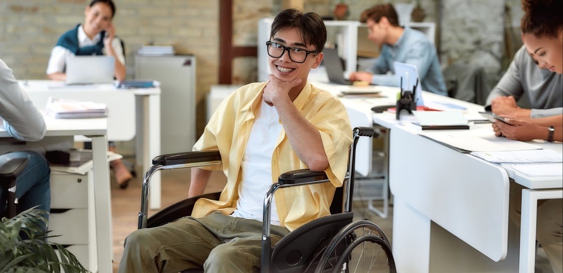 A person in a wheelchair smiling at the camera, while seated in a modern office environment with other individuals working in the background.