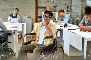 A person in a wheelchair smiling at the camera, while seated in a modern office environment with other individuals working in the background.